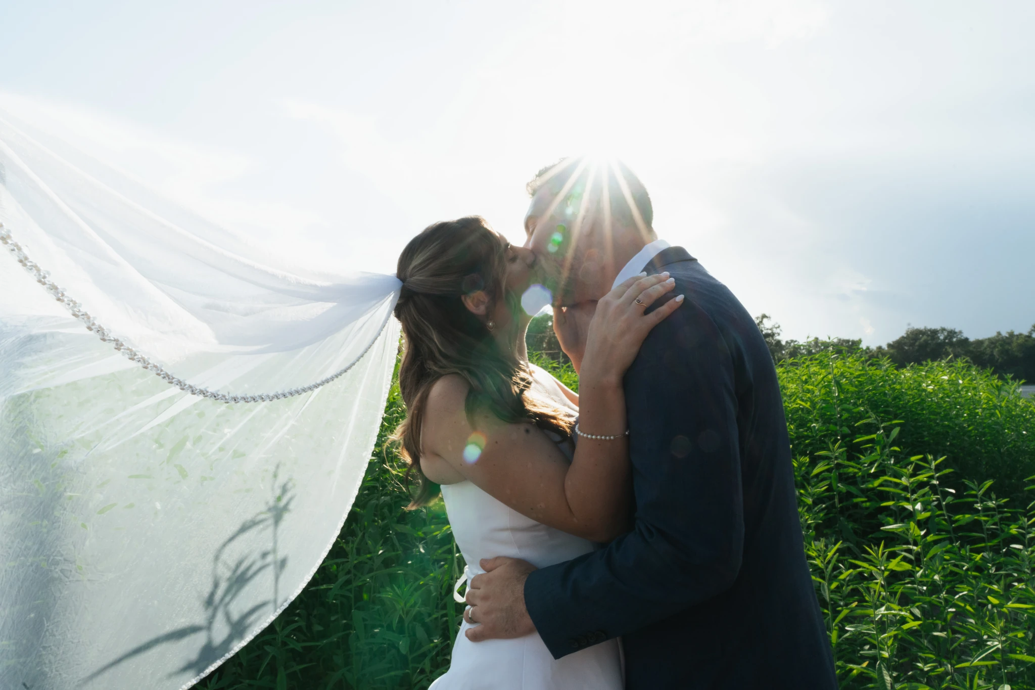 A bride and groom kiss into the sun as her veil streams out in the wind