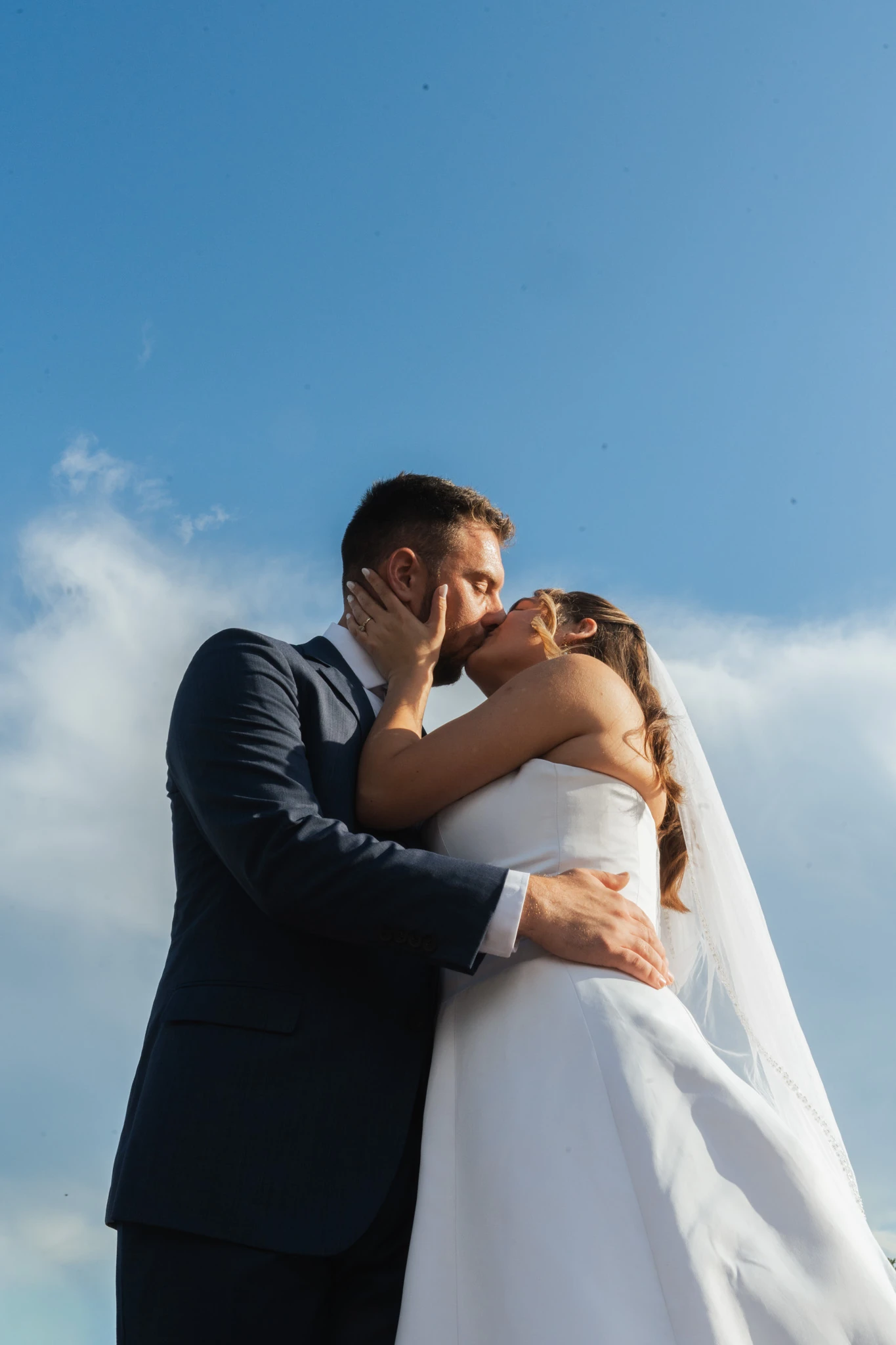 The couple kiss against a blue sky, her veil lifting behind her