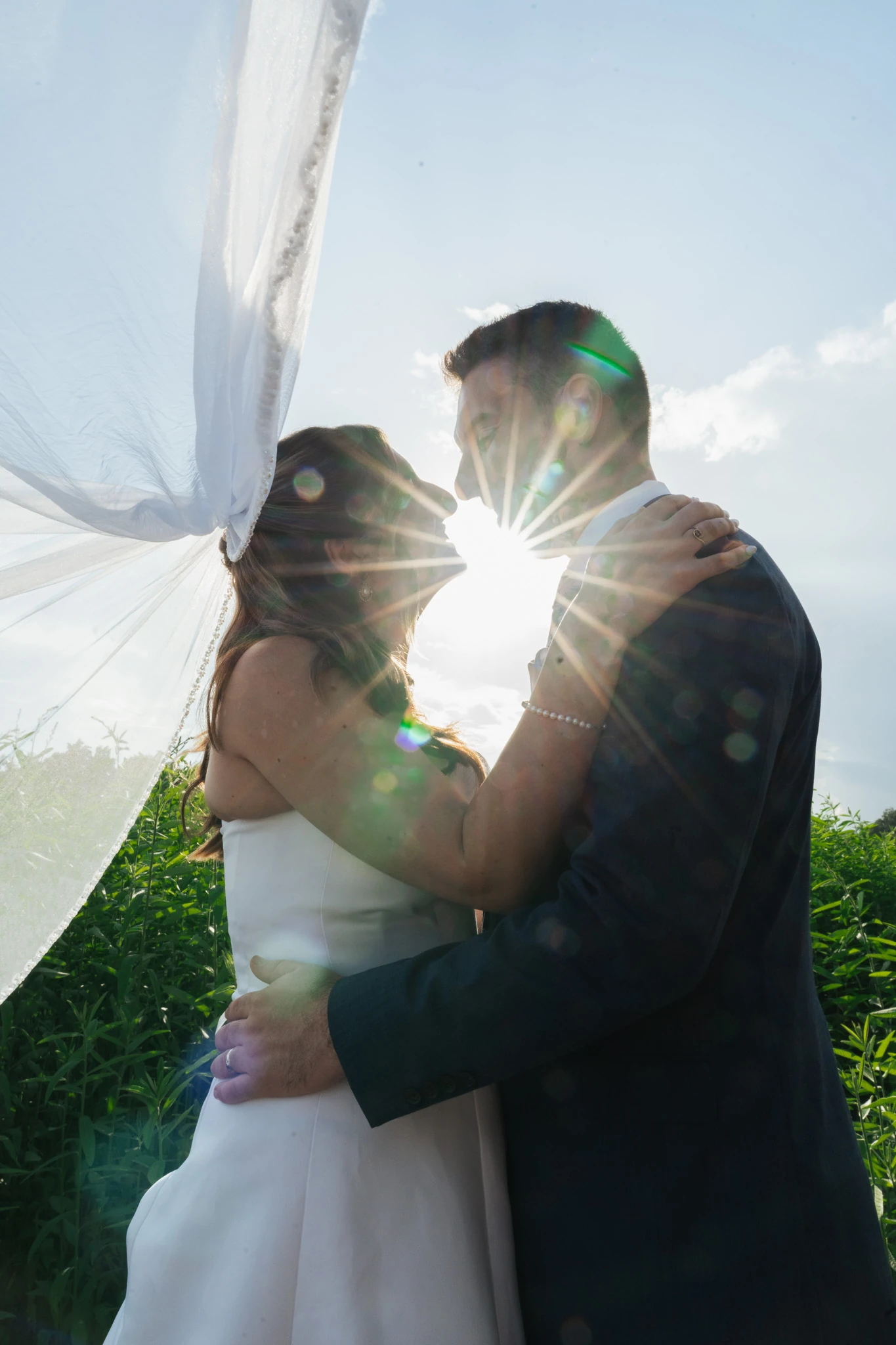 The sun flares between the couple as the veil lifts in the wind