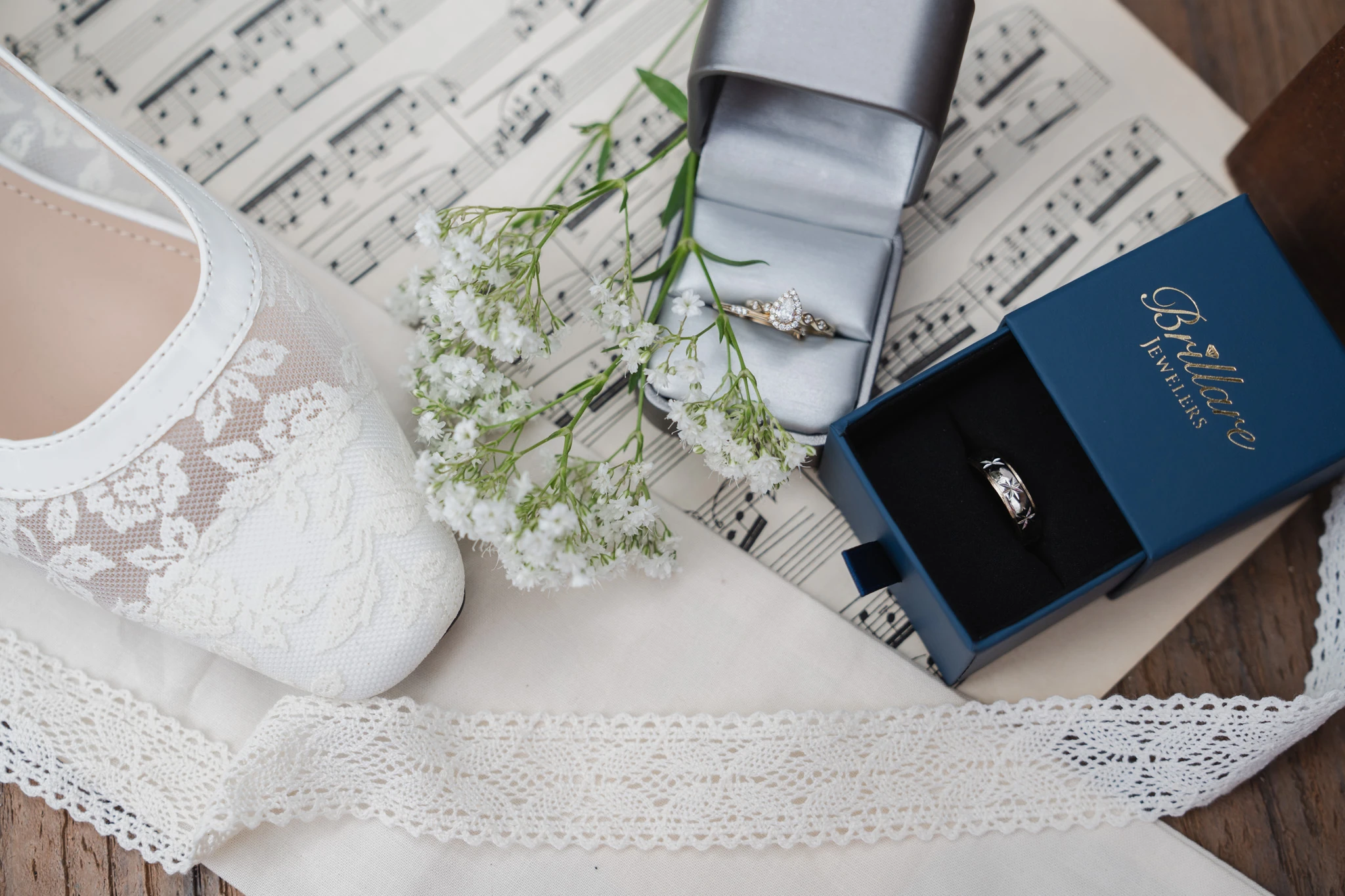 Two wedding rings in their boxes beside lace bridal shoes and baby's breath on sheet music